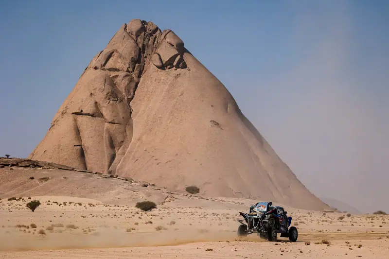 Ein SSV fährt bei der Dakar 2021 durch die Wüste vor einem riesigen Felsen